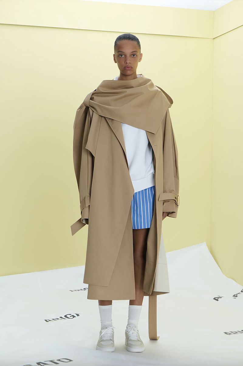 A person stands in a photo studio wearing a tan trench coat, a white sweatshirt, and blue-striped shorts. The background is a solid yellow.