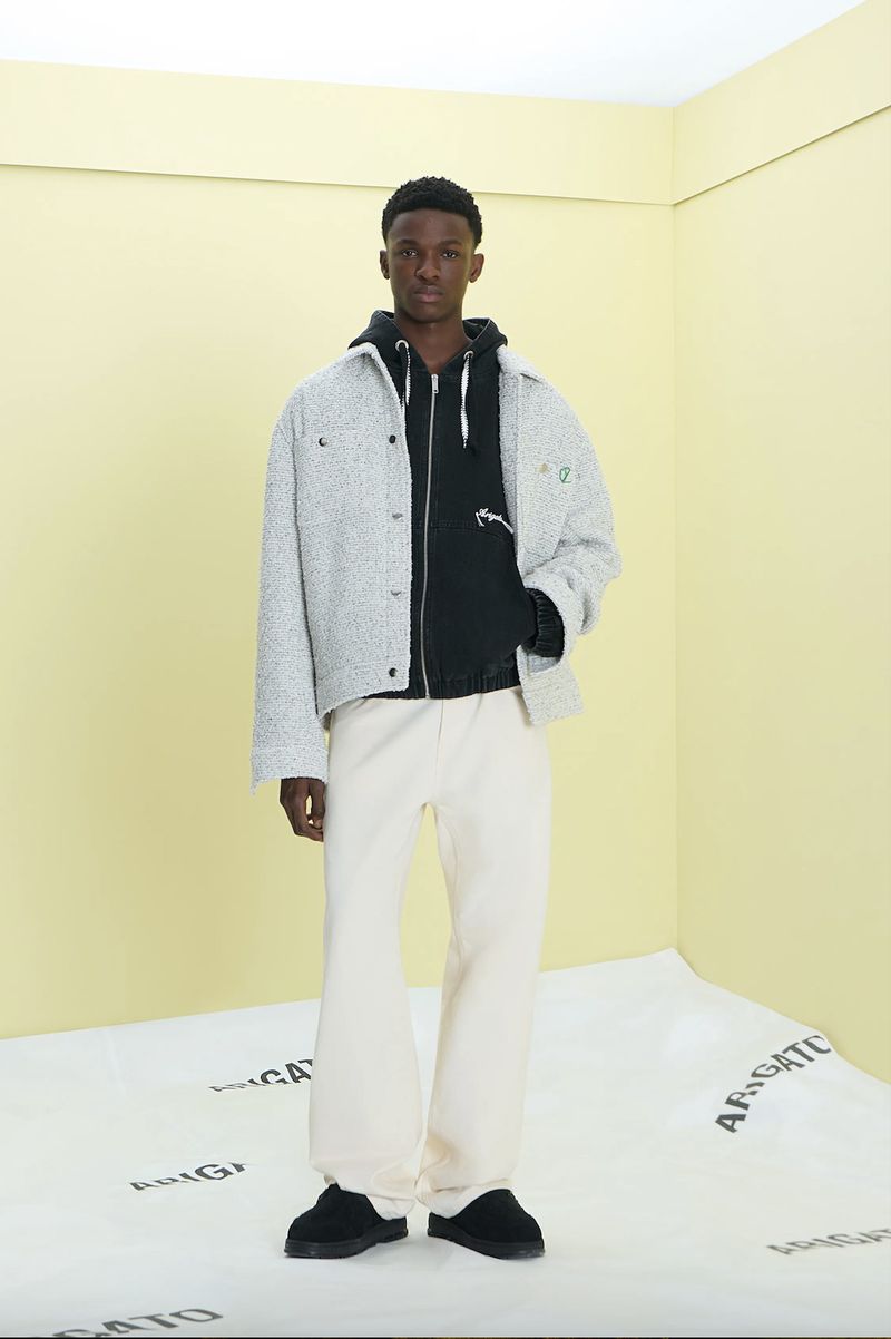 Young man in streetwear fashion: grey overshirt, black hoodie, and baggy off-white trousers, poses in minimalist photo studio with repeating text.