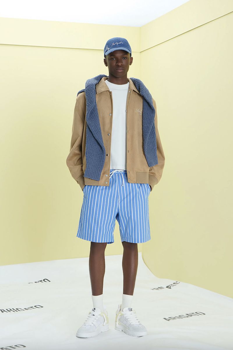 Model wearing a tan jacket, blue striped shorts, shoulder-draped knit sweater, and white sneakers against a pale yellow studio set.