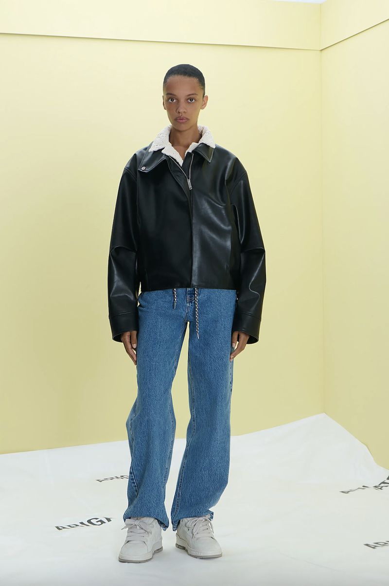 Fashion model poses indoors against a two-tone backdrop wearing a black leather jacket with a white shearling collar, and blue jeans.