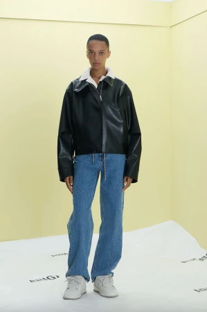 Fashion model poses indoors against a two-tone backdrop wearing a black leather jacket with a white shearling collar, and blue jeans.
