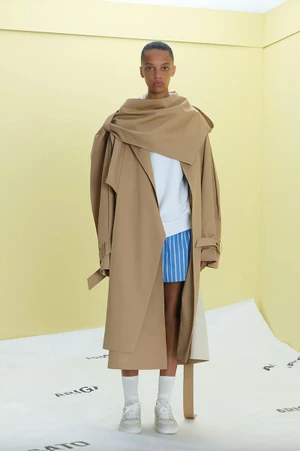 Model wearing a large camel trench coat, white sweatshirt, and striped shorts against a pale yellow studio background.