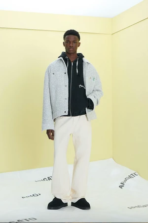Model wearing a light grey textured jacket over a black hoodie and cream trousers against a yellow studio backdrop.