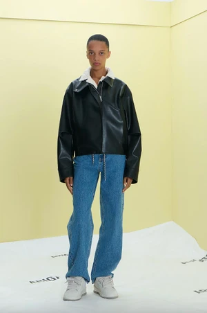 Model wearing an oversized black leather jacket, wide-leg blue jeans, and white sneakers against a pale yellow studio backdrop.