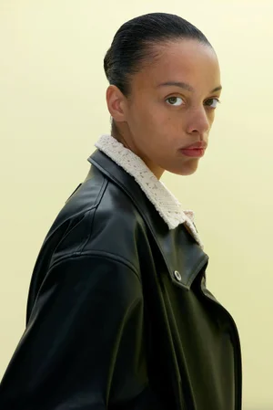 Portrait of a model wearing a black jacket with a white textured collar against a plain pale background