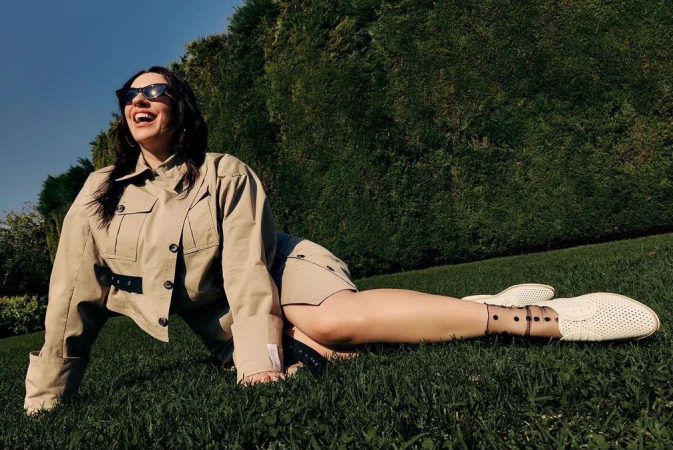 A woman lies on a lawn wearing a tan jacket, shorts, sunglasses, and white shoes, laughing against a background of grass and greenery.