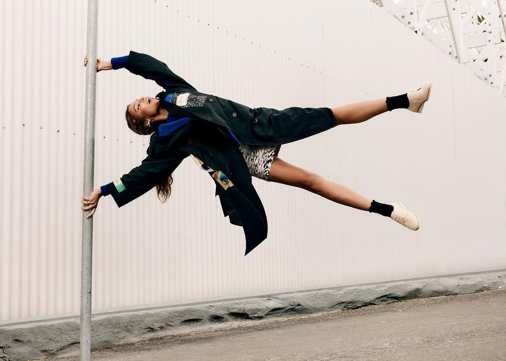Woman in black coat and patterned dress strikes an acrobatic pose outdoors, suspended and holding a pole, with white wall backdrop.
