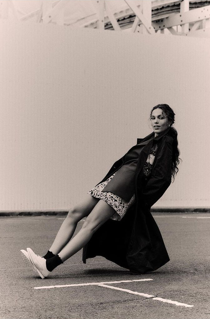 Monochrome fashion shot of a woman in a black coat and lace-trimmed dress, angled pose on asphalt, white sneakers, against a neutral background.