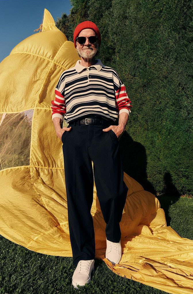 Mature man in red hat and sunglasses stands in front of a yellow backdrop in outdoor setting, wearing a striped collared shirt.