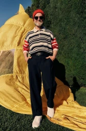 Elderly man in a red beanie and striped polo shirt standing outdoors with a large yellow inflatable prop.