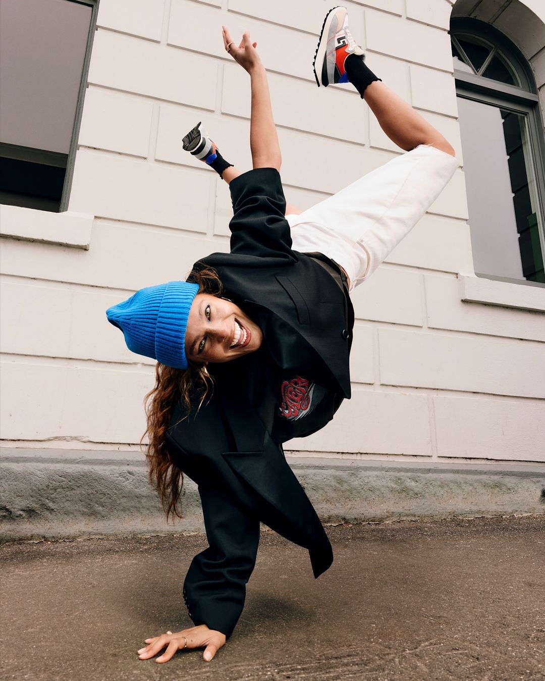Woman in a blue beanie and black jacket doing a handstand outdoors against a pale tiled building exterior