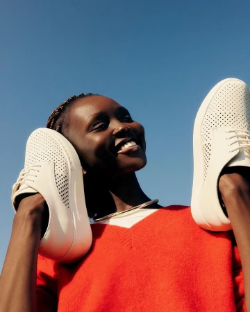 Smiling Black woman wearing a bright red sweater holding two white perforated shoes against a clear blue sky