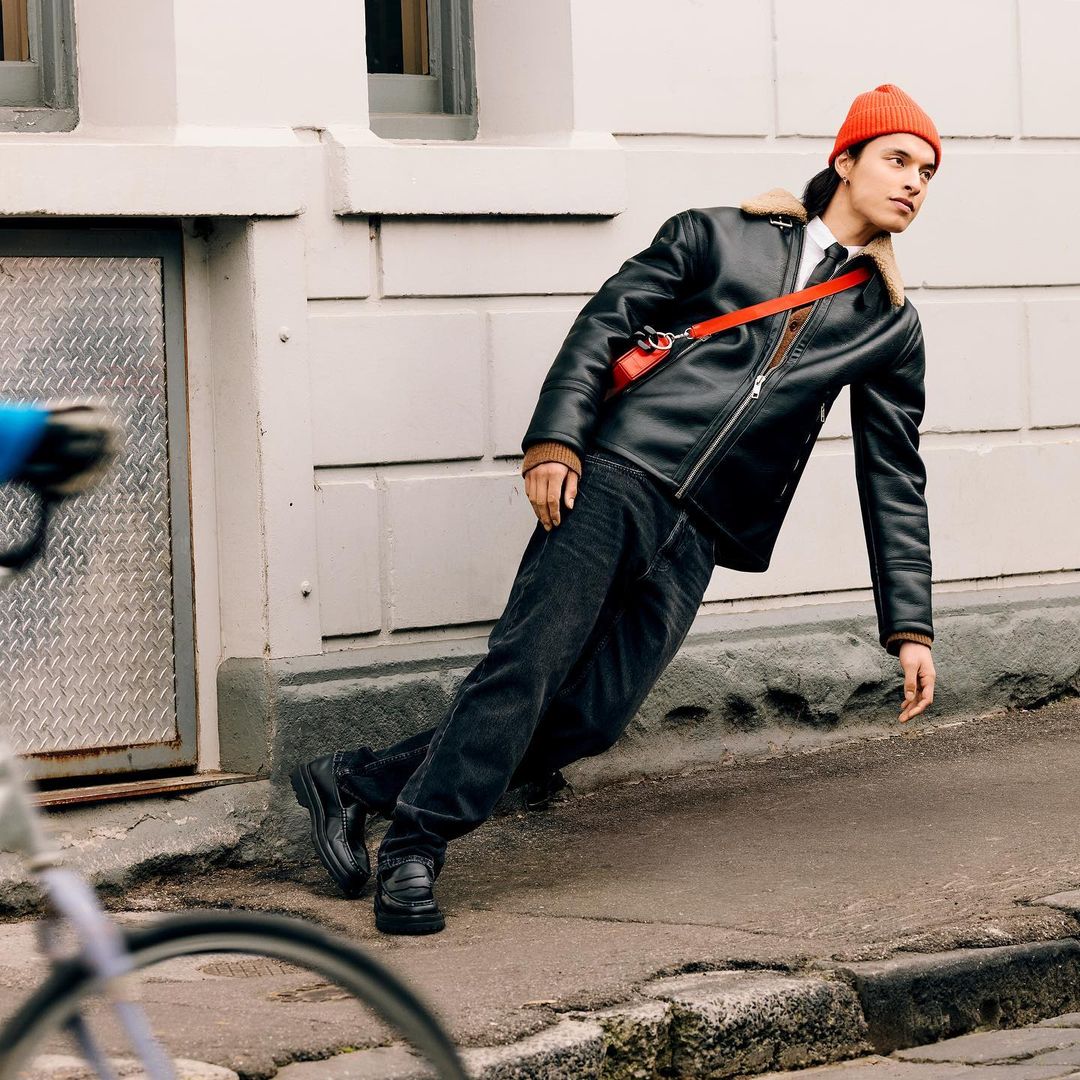 Man in black shearling jacket and orange beanie leans against building wall with bicycle wheel visible in the lower left foreground.