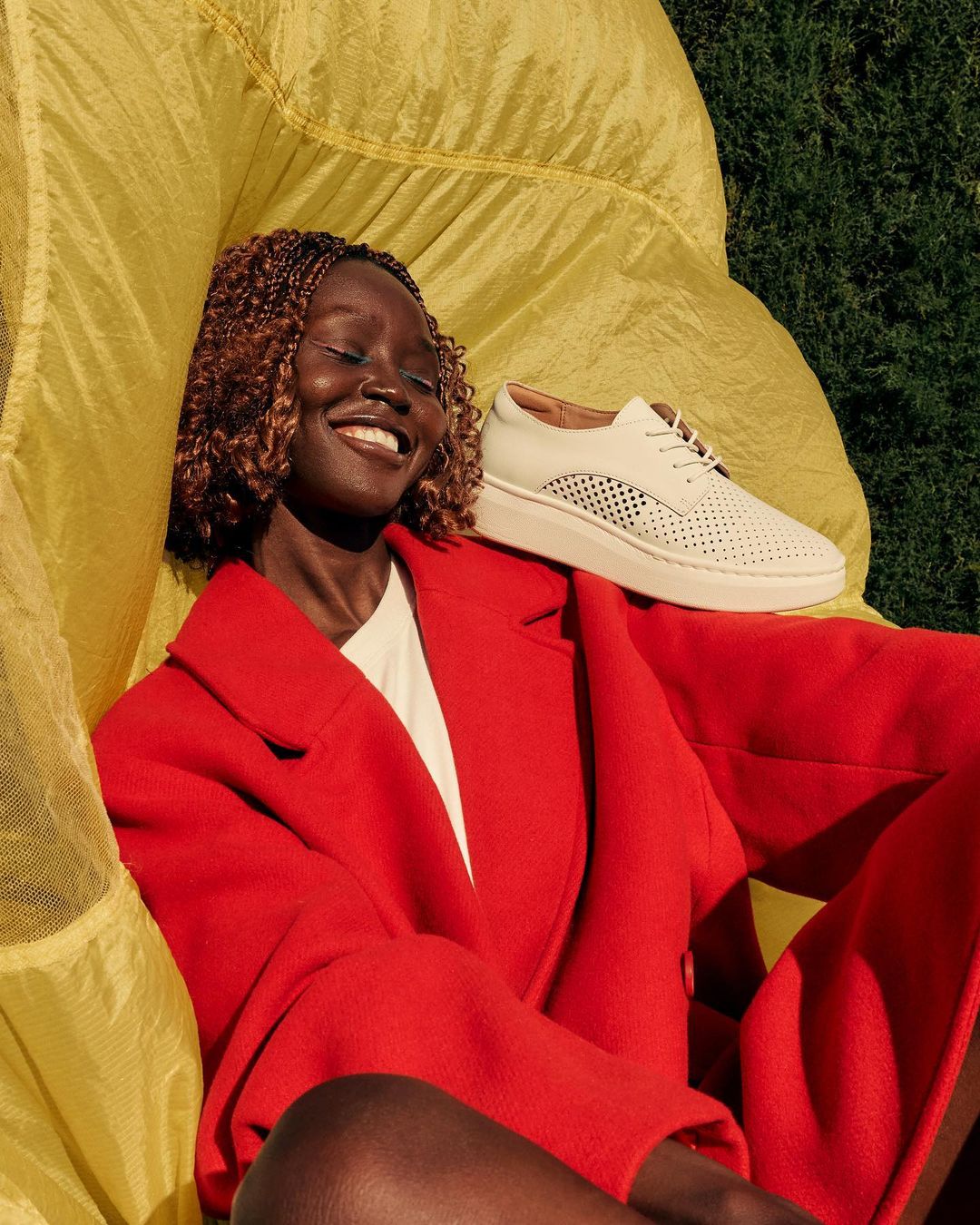 Smiling Black woman poses with a white sneaker on her shoulder, wearing a red coat against a yellow fabric and green foliage background.