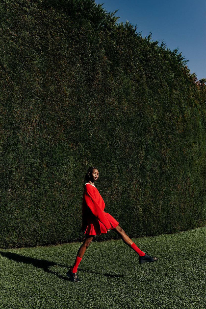 Adult female in a red dress and tights walks in front of a tall green hedge on a grassy lawn under a clear blue sky on a sunny day.