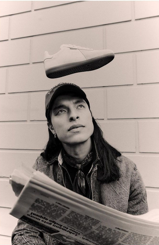 A man with long hair and a baseball cap holds a newspaper as a shoe floats above his head against a light brick wall background.