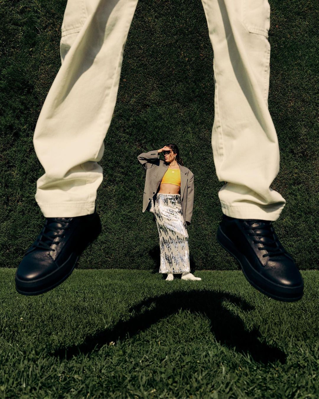 Low angle shot of a person jumping in white trousers and dark sneakers over grass, with another model standing in the distance.