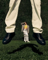 Low angle shot of a person jumping in white trousers and dark sneakers over grass, with another model standing in the distance.