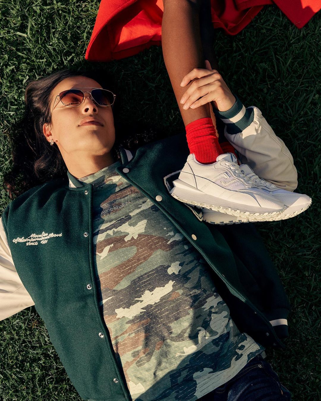 Two people lying on green grass in natural daylight. One person is wearing a green and white jacket and aviator sunglasses.