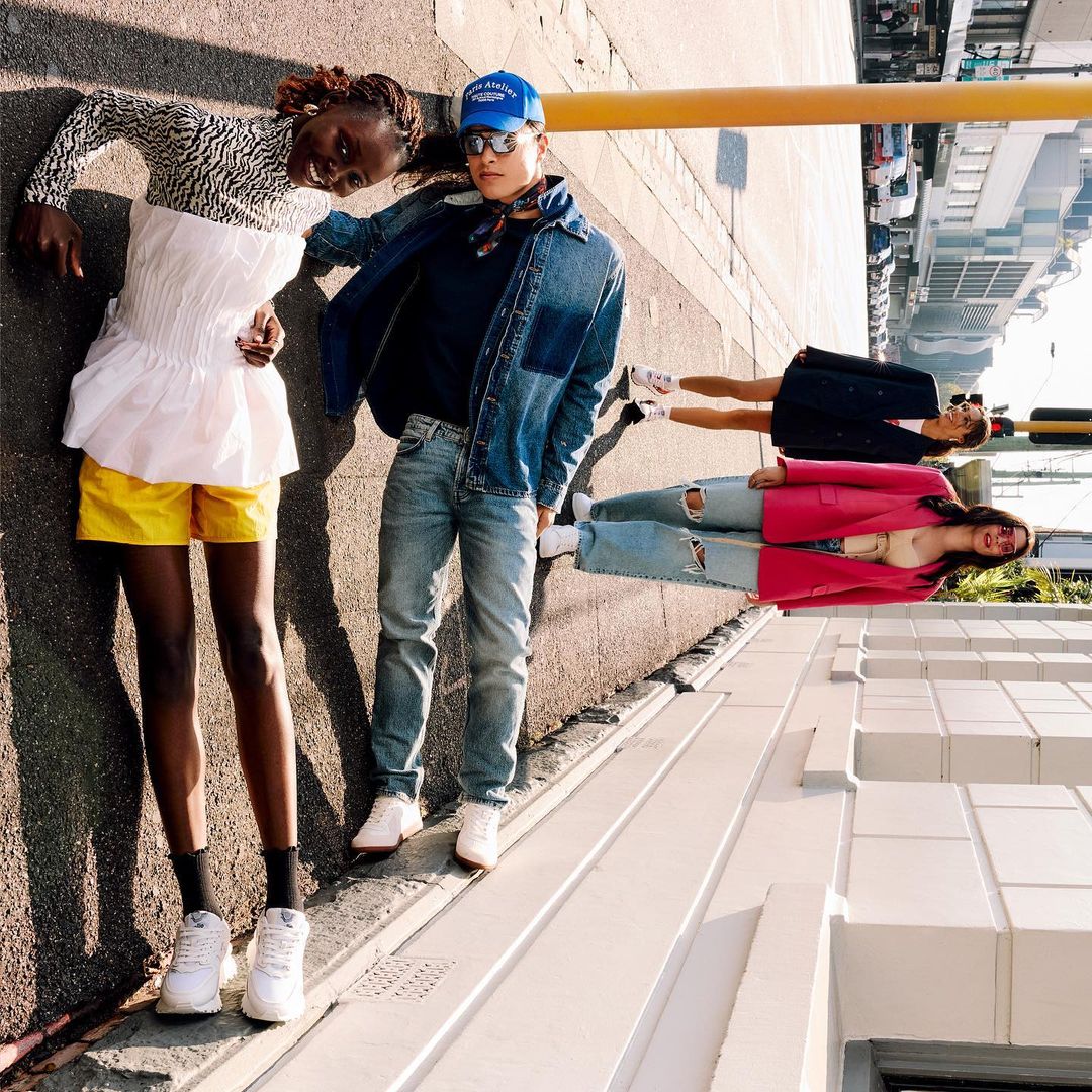Three stylish people pose outdoors with two figures rotated on their sides; fashion editorial shot on a sunny urban street with bright colors.