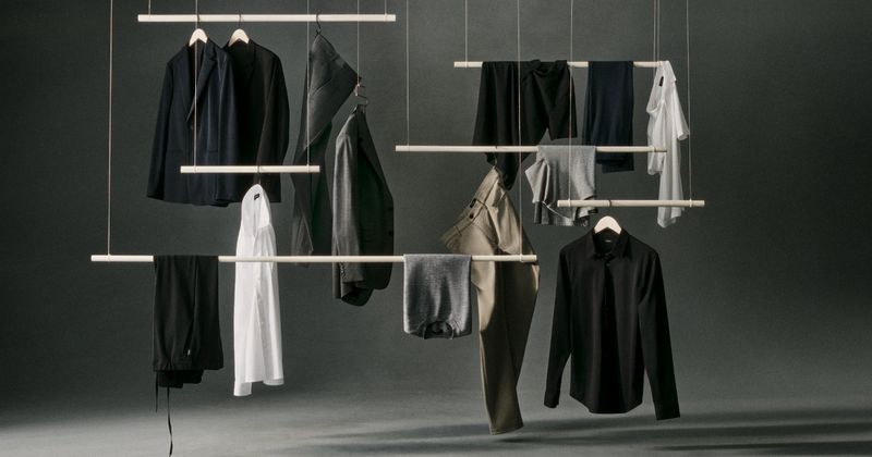 Assortment of menswear including jackets, shirts, and pants hanging on light wood racks against a dark neutral background in studio lighting.