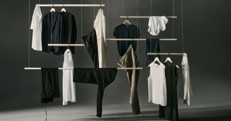 Clothing collection display featuring a curated assortment of garments suspended on wooden racks against a minimalist grey background.