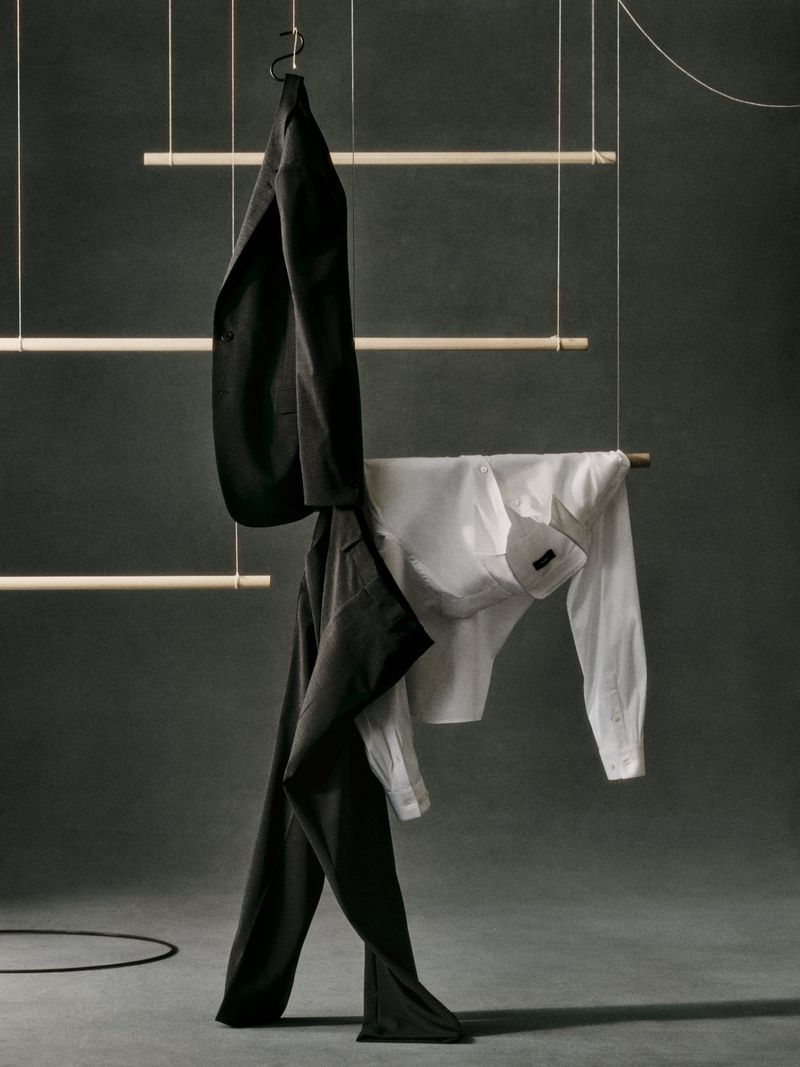 Studio shot of a black suit and a white dress shirt hanging from a wooden horizontal ladder, showcasing contemporary fashion.
