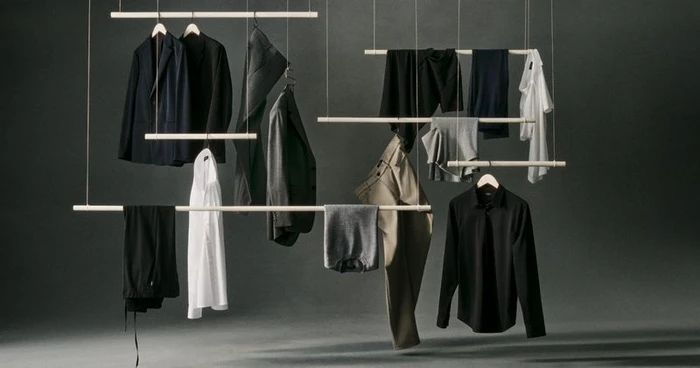 Suspended wooden rods hold hanging garments and folded apparel against a dark grey textured studio background.