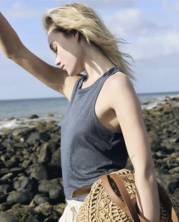 Woman posing outdoors on a rocky shore wearing a navy tank top, white bottoms, and carrying a straw bag with leather handles.