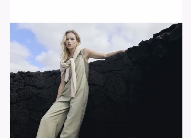 Fashion editorial shot of a blonde woman in a light green jumpsuit posing against a dark volcanic rock formation under a cloudy sky.