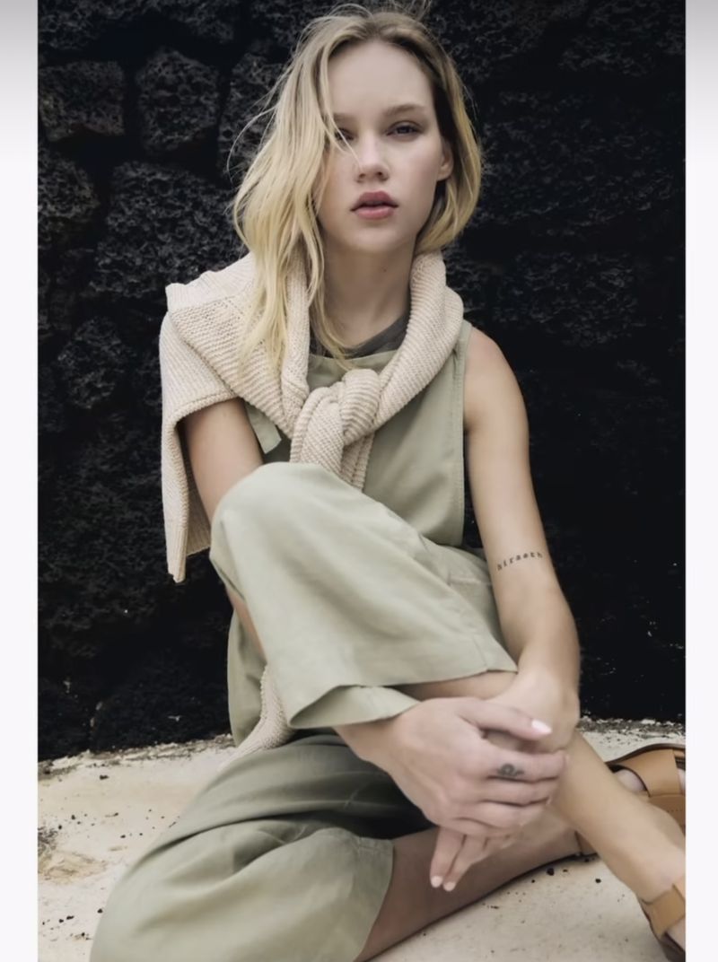 A fair-skinned woman with blonde hair sits outdoors, wearing a sage green jumpsuit and cream sweater, next to a dark stone wall.