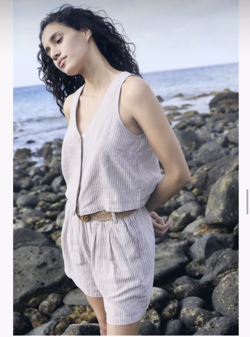 Young woman in a pinstripe linen two-piece outfit with a woven belt standing on a rocky beach with the ocean in the background.