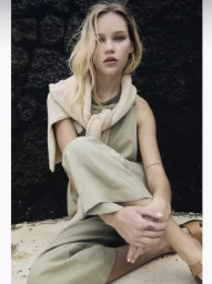 Model Evelina Sakalauskaite seated against a dark volcanic rock wall wearing a sage green linen outfit and cream knit sweater.