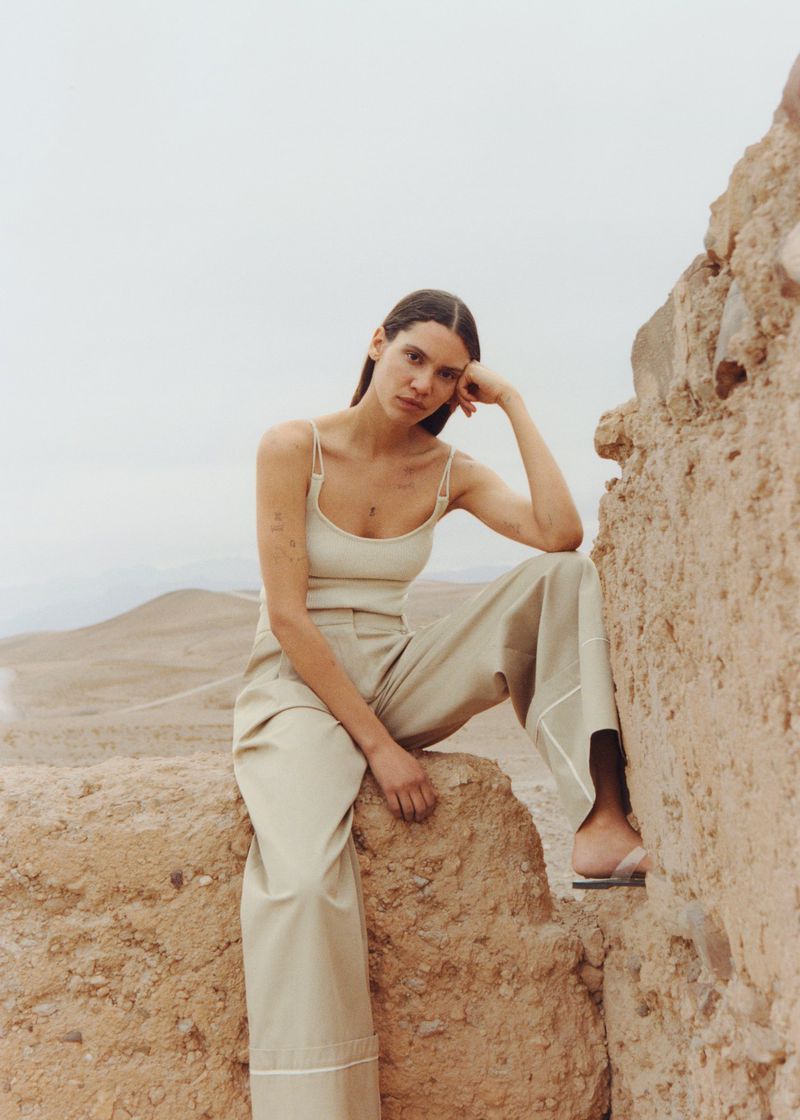 A young woman in neutral-colored clothing is sitting on a rocky desert overlook on a cloudy day in a full outdoor shot.