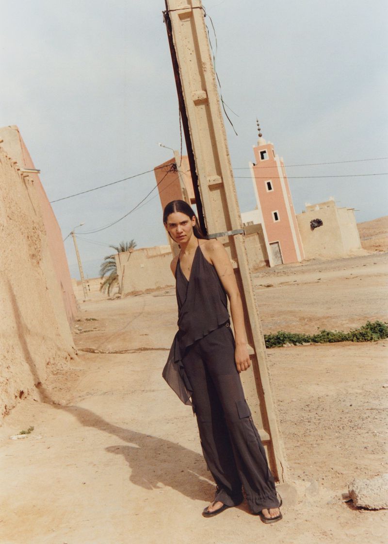 A young woman in a dark outfit poses on a dusty street near a light pole, with a pale-pink mosque visible in the background under a blue sky.