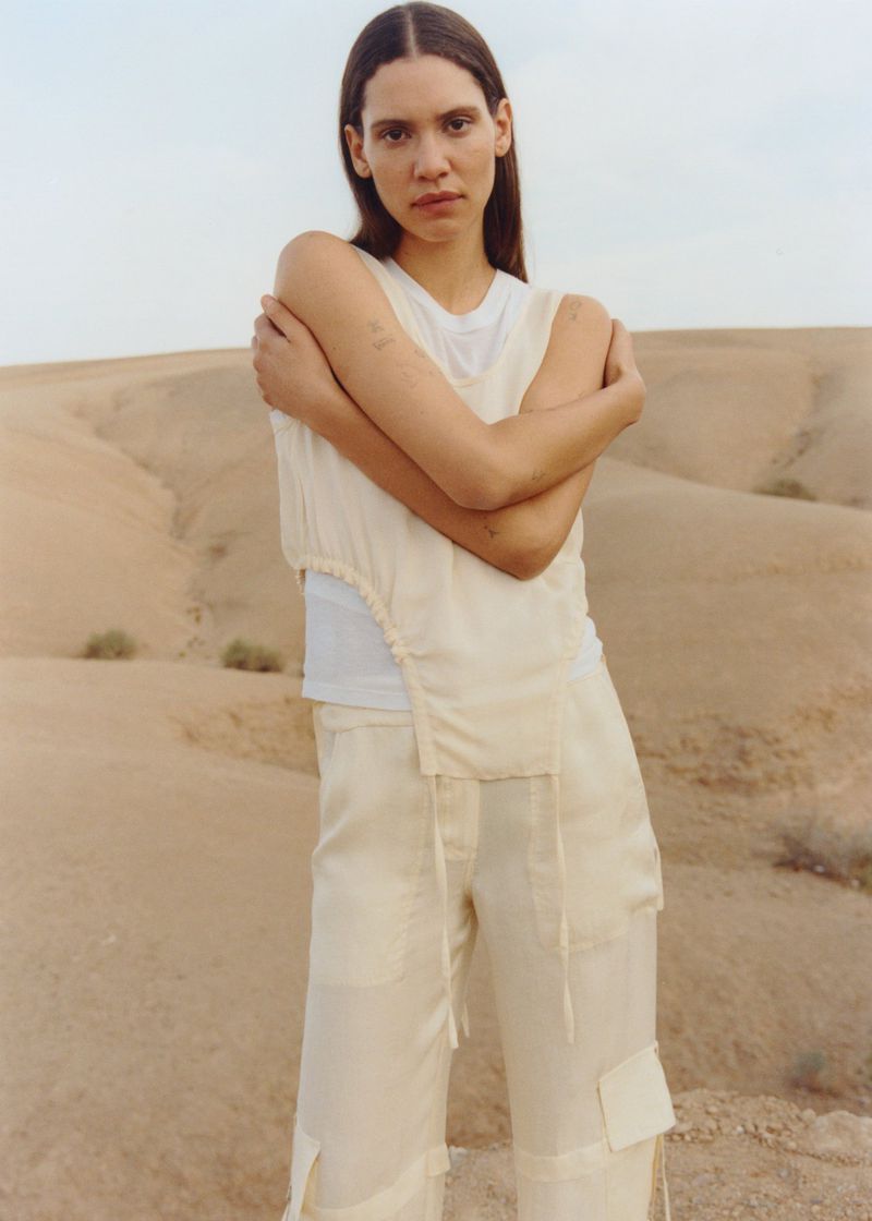 A young woman stands in a desert landscape, wearing a light-colored sleeveless shirt and pants, with rolling sand dunes in the background.