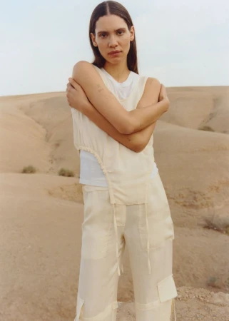 A young woman stands in a desert landscape, wearing a light-colored sleeveless shirt and pants, with rolling sand dunes in the background.