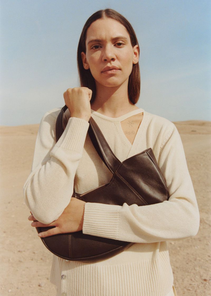A woman in a cream sweater poses outdoors with a dark brown leather handbag in a desert landscape under a clear blue sky.