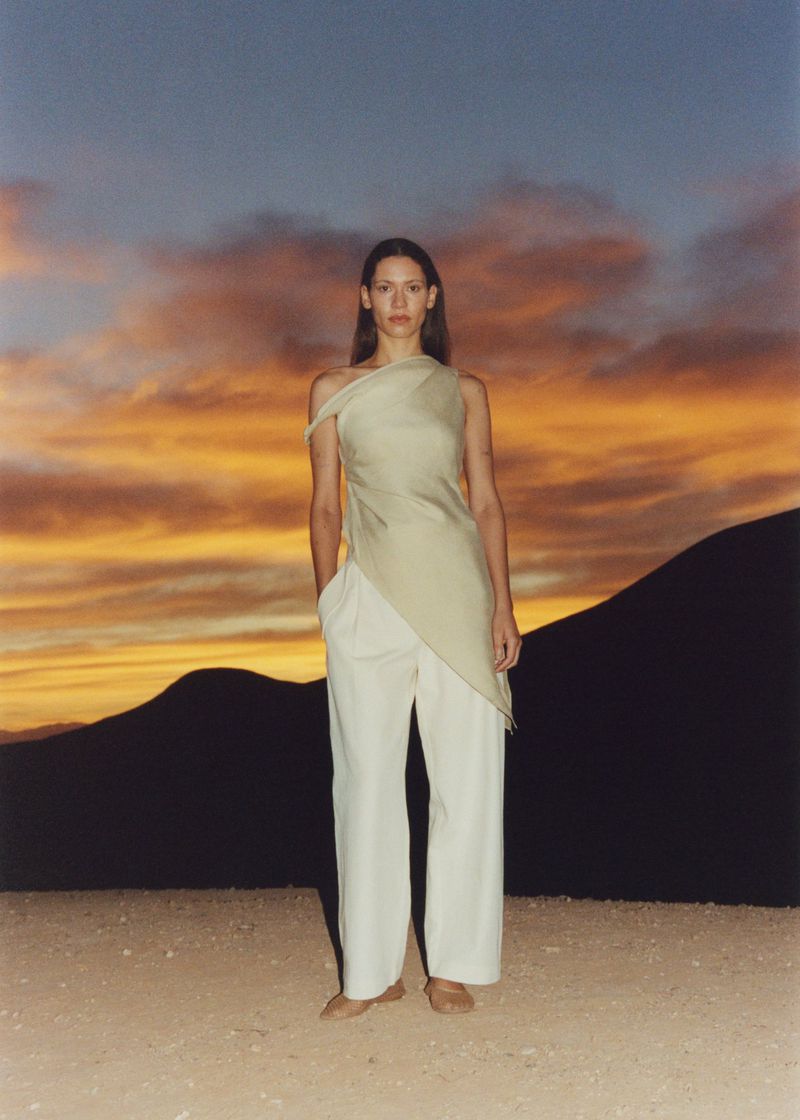 A woman poses in an asymmetrical top and wide-legged pants against a backdrop of silhouetted mountains and a dusky, orange-streaked sky at dusk.