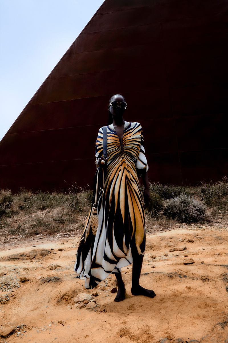Dark-skinned person stands in a tiger-striped dress with a brown pyramid and blue sky in the background, holding a shoulder bag.
