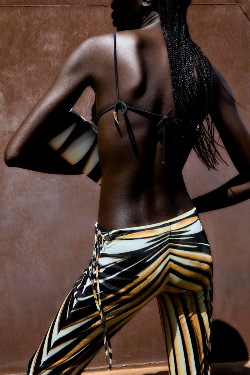 Back view of a Black model wearing animal print pants and top, carrying a zebra print purse against a solid brown background in natural light.