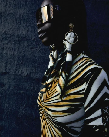 Fashion portrait of a stylish dark-skinned woman wearing sunglasses, patterned top, scarf, and hoop earrings against a dark wall.