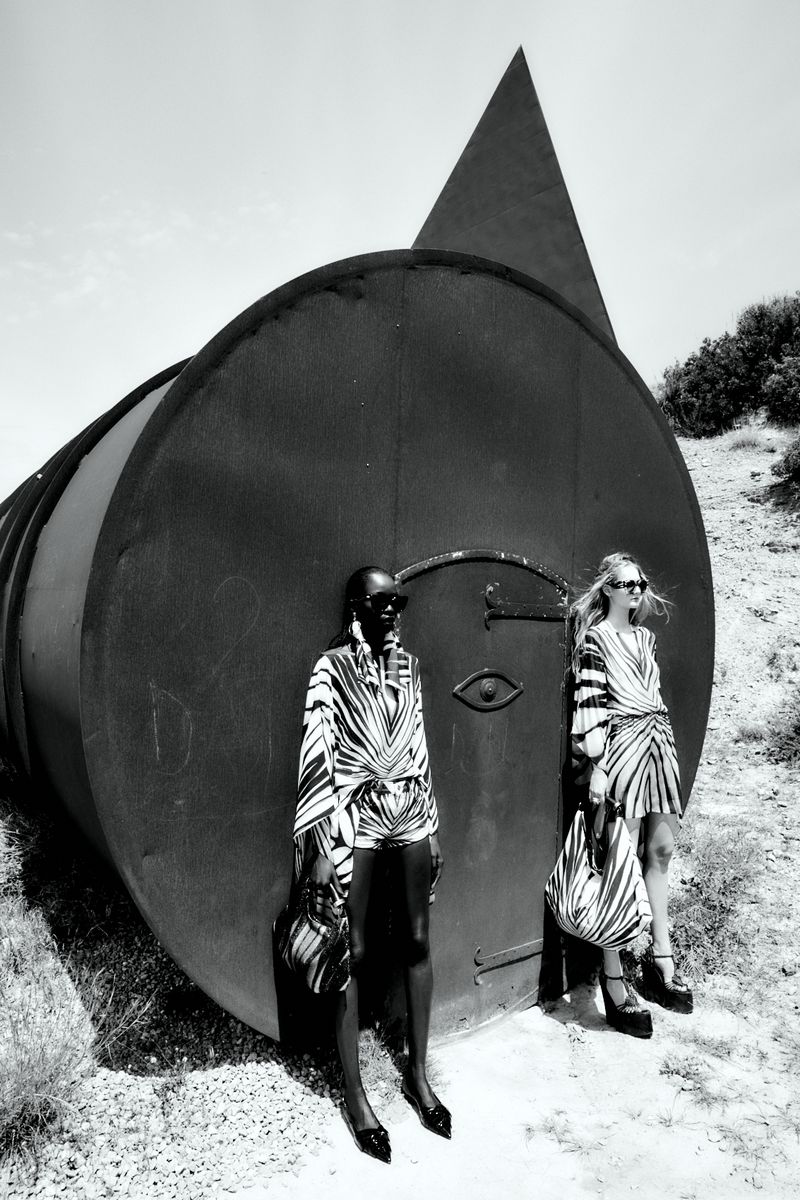 Two fashion models pose in patterned outfits beside a large metal cylinder in a black and white outdoor fashion shot.