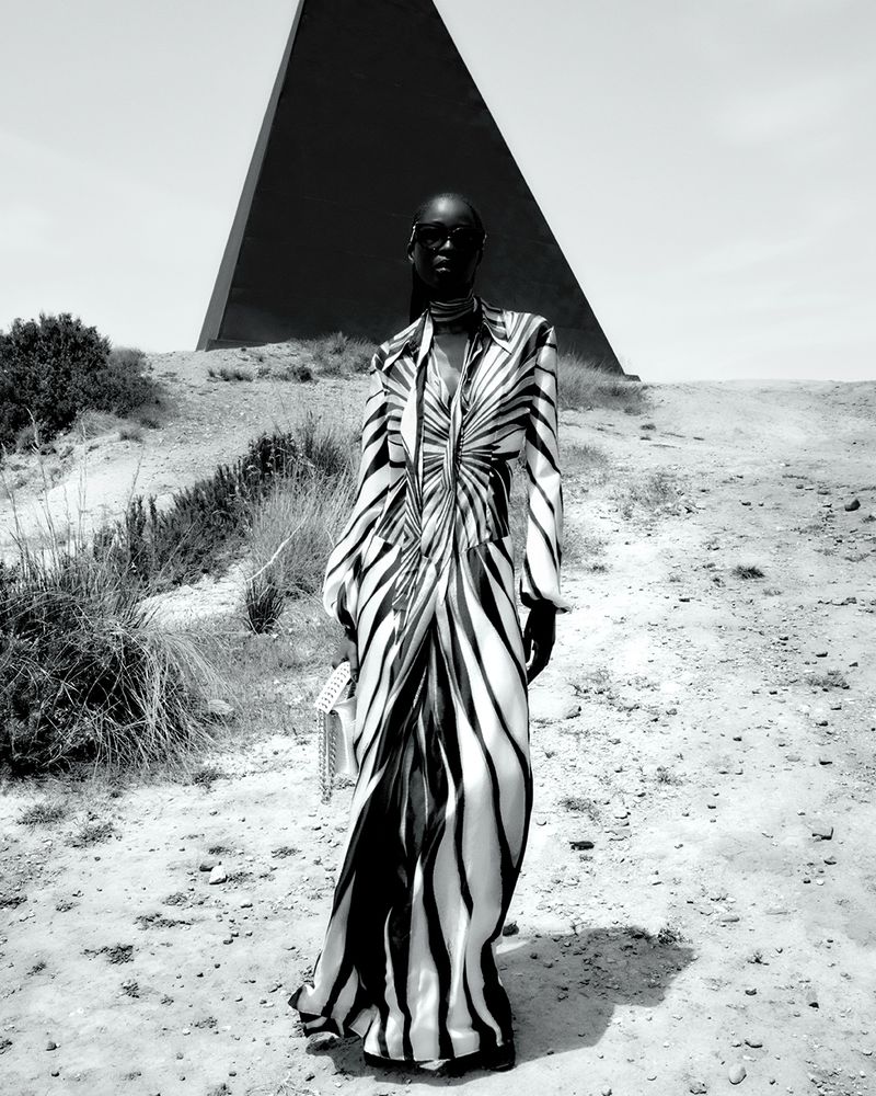 Fashionable Black woman in a zebra-striped dress and sunglasses stands in a desert landscape with a geometric pyramid structure.