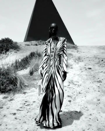 Fashionable Black woman in a zebra-striped dress and sunglasses stands in a desert landscape with a geometric pyramid structure.