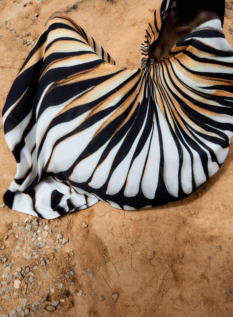A woman in a patterned dress sits on a sandy surface, creating a visually striking composition with contrasting textures and colors.