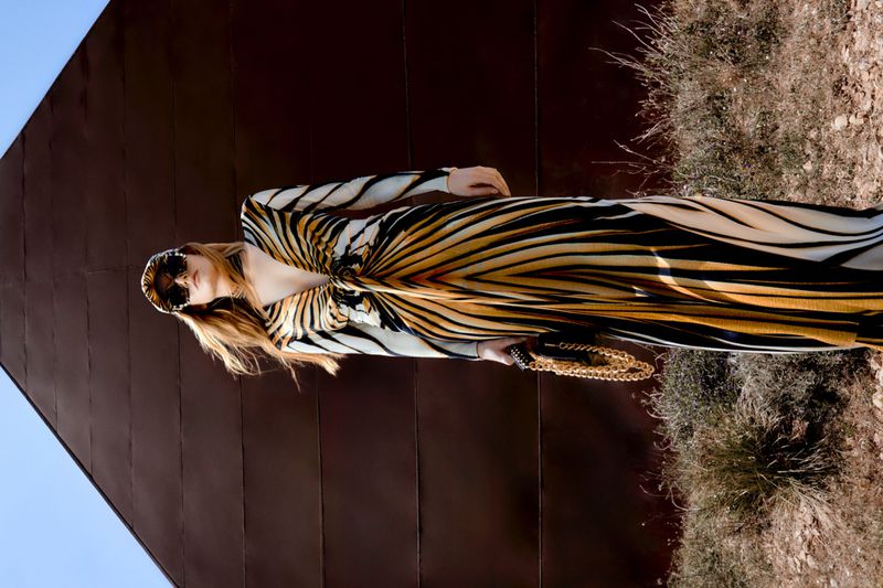 Woman in a black, tan, and white tiger-striped dress, headscarf, and sunglasses poses in front of a brown wall and dry foliage outdoors.