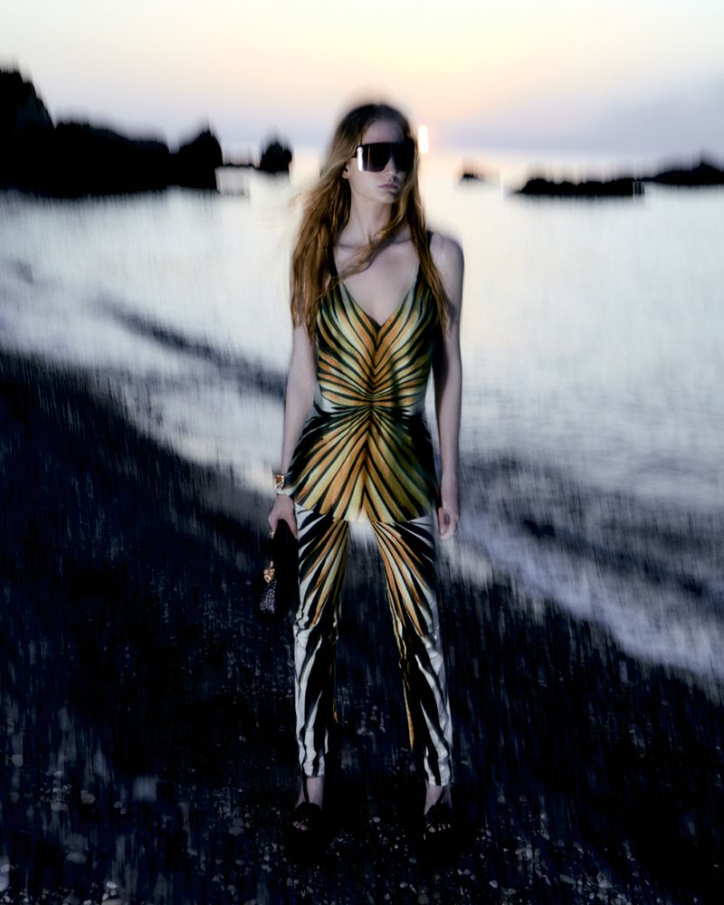 Adult woman in patterned jumpsuit on a beach at dusk, wearing sunglasses and carrying a clutch.