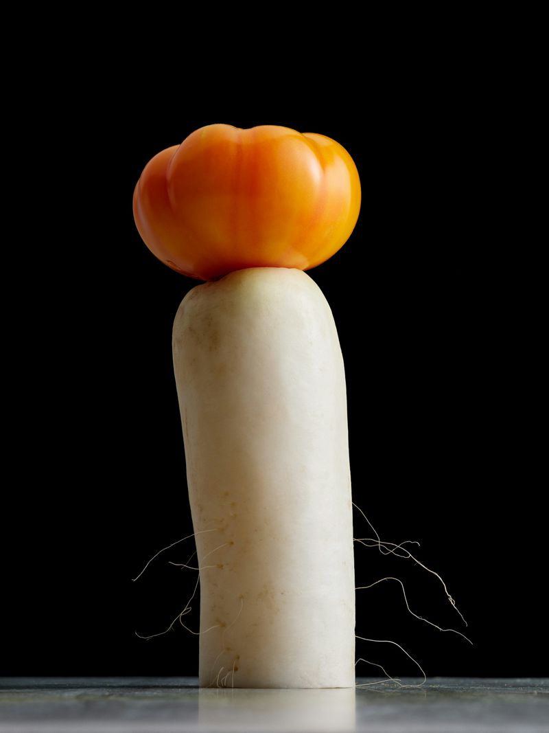 Still life photograph featuring a white daikon radish topped with an orange heirloom tomato against a dark background, creating a minimalist food composition.