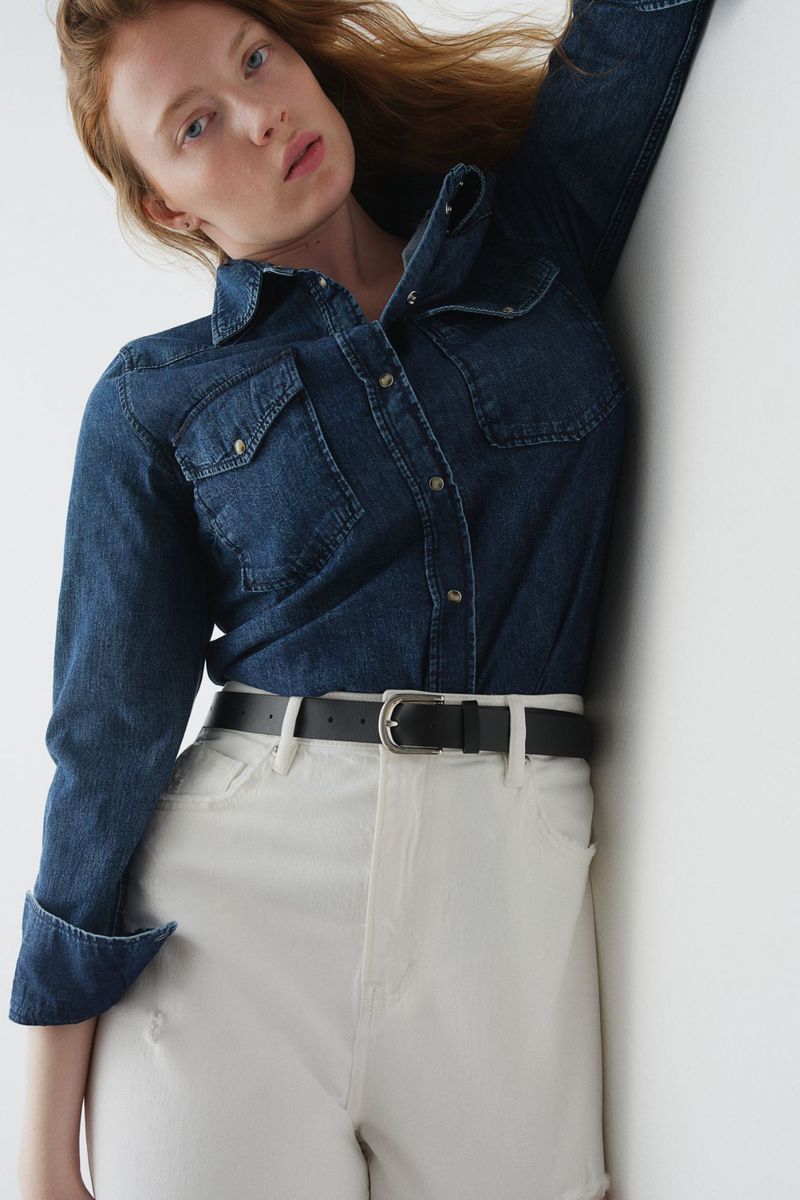 A fashion shot of a young adult woman with red hair wearing a denim shirt and white jeans indoors against a white wall.